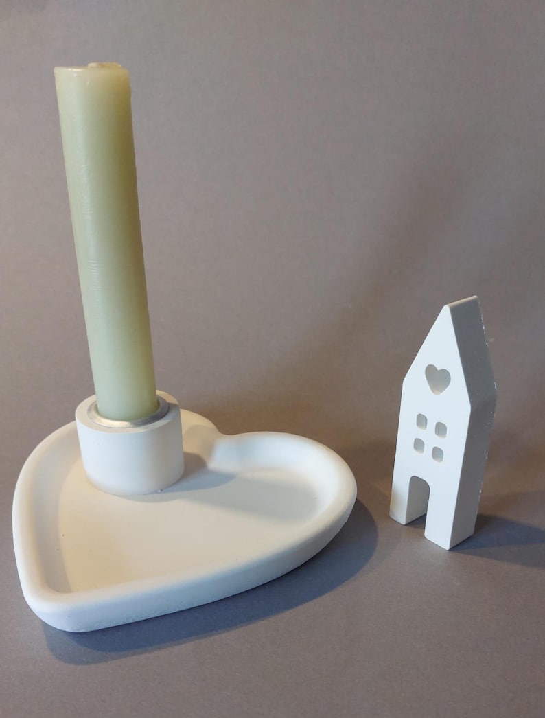 Könnte beinhalten: Ein weißer, herzförmiger Kerzenhalter mit einer hohen, hellgrünen Kerze. Daneben eine kleine, weiße hausförmige Dekoration mit einem Herz-Ausschnitt und Fensterdetails. Die Gegenstände stehen auf einer grauen Oberfläche.