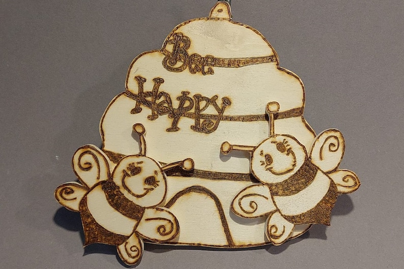Könnte beinhalten: Ein Holz-Bienenstock-Anhänger mit der Aufschrift "Be Happy". Zwei Bienen mit schwarzen und weißen Streifen fliegen um den Bienenstock herum.