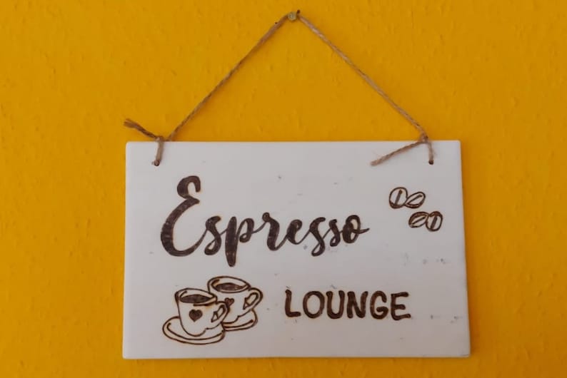 Könnte beinhalten: Ein rechteckiges weißes Schild mit den Worten "Espresso LOUNGE" in handschriftlicher Schrift. Das Schild zeigt auch Illustrationen von Kaffeebohnen und zwei Kaffeetassen. Das Schild hängt an einer Schnur vor einem gelben Hintergrund.