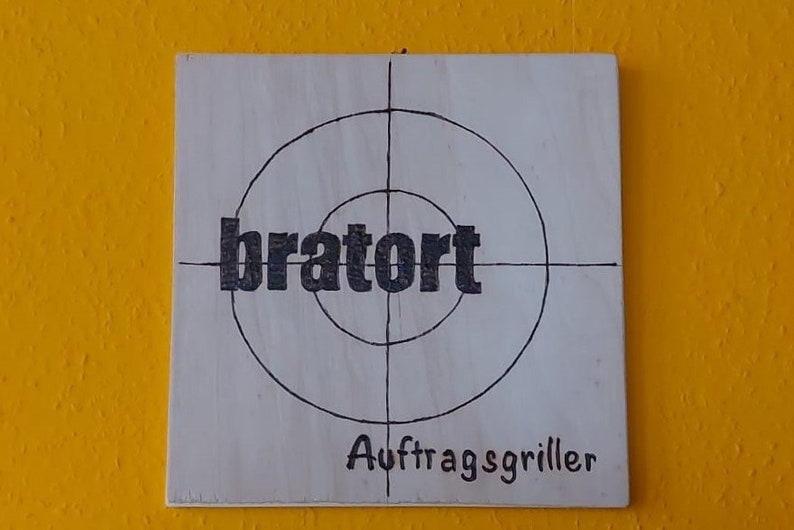 Könnte beinhalten: Ein weißes Holzschild mit einer schwarzen Zielscheibe darauf. Das Wort "bratort" ist in der Mitte der Zielscheibe geschrieben. Unter der Zielscheibe stehen die Worte "Auftragsgriller" in Schwarz.
