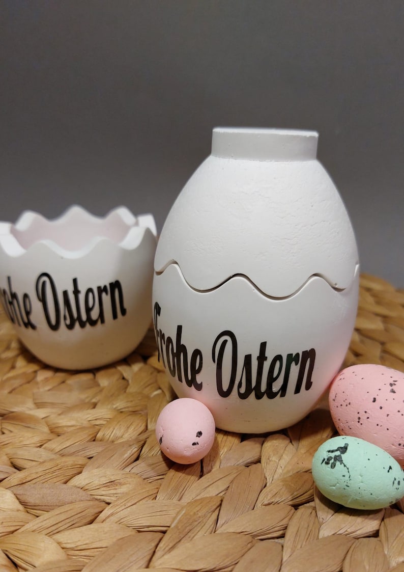 Könnte beinhalten: Weißer eiförmiger dekorativer Behälter mit Deckel und ein passender eierschalenförmiger Behälter, beide mit dem Text "Frohe Ostern" in schwarzer Schrift. Mehrere gesprenkelte Pastell-Eier sind ebenfalls vorhanden, auf einer gewebten Oberfläche.