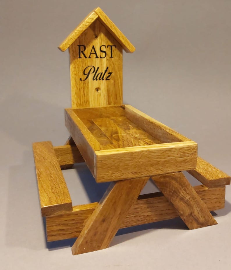 Könnte beinhalten: Ein hölzerner Vogelhäuschen in Form einer Picknickbank mit einem kleinen Häuschen oben drauf. Das Häuschen hat die Worte "Rast Platz" eingraviert.