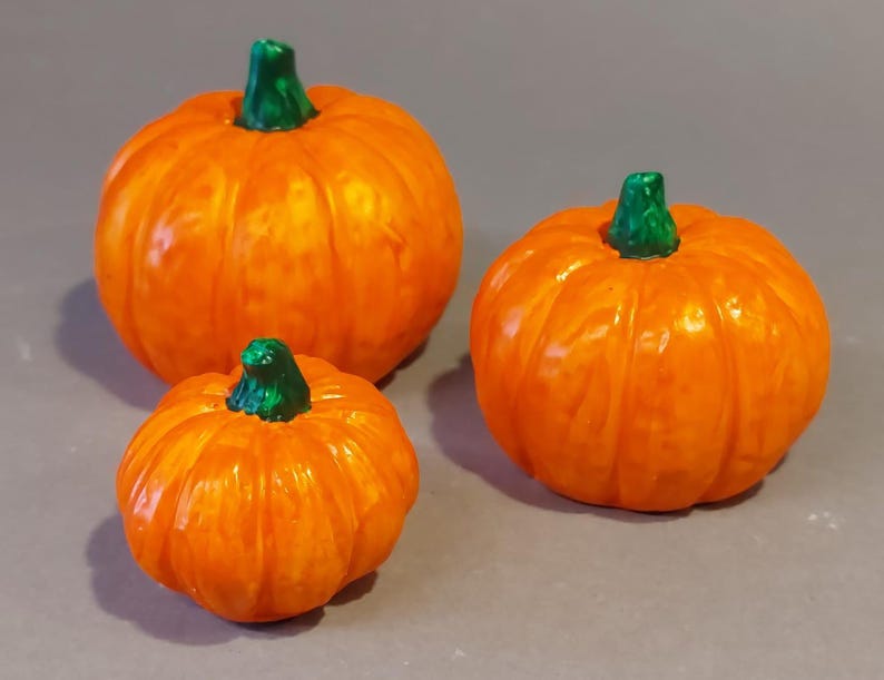 Könnte beinhalten: Drei orangefarbene Kürbisse mit grünen Stielen sind auf einer grauen Oberfläche angeordnet. Die Kürbisse sind unterschiedlich groß, der größte hinten und der kleinste vorne. Die Kürbisse haben eine glänzende Oberfläche.