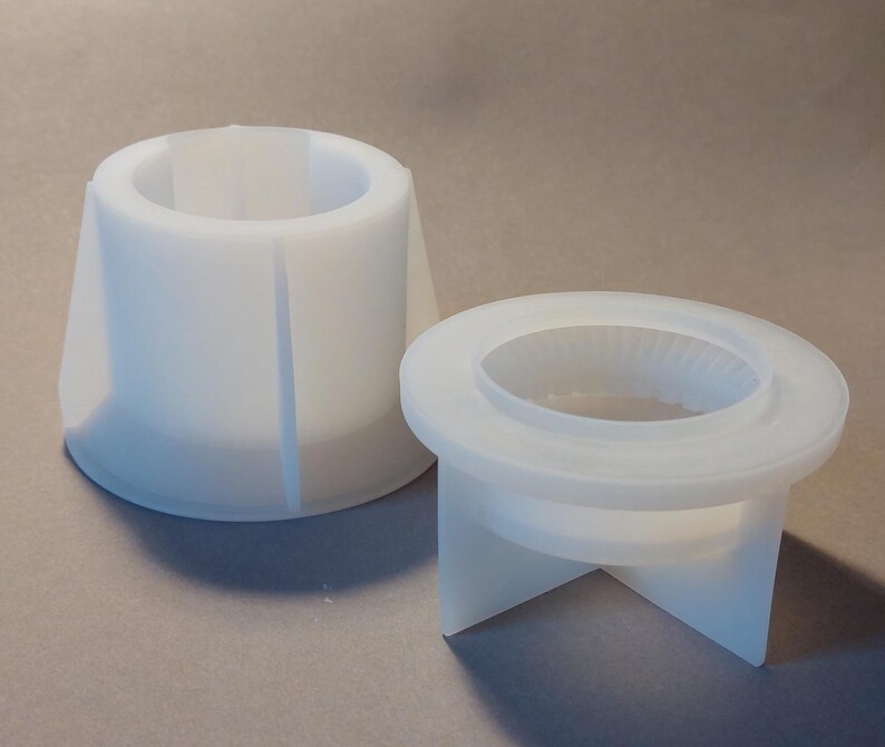 Könnte beinhalten: Zwei weiße Silikonformen zum Basteln. Eine ist eine hohe, konische Form mit einer breiten Öffnung und vertikalen Aussparungen. Die andere ist eine kurze, kreisförmige Form mit einer zentralen Öffnung und einem dreibeinigen Sockel.