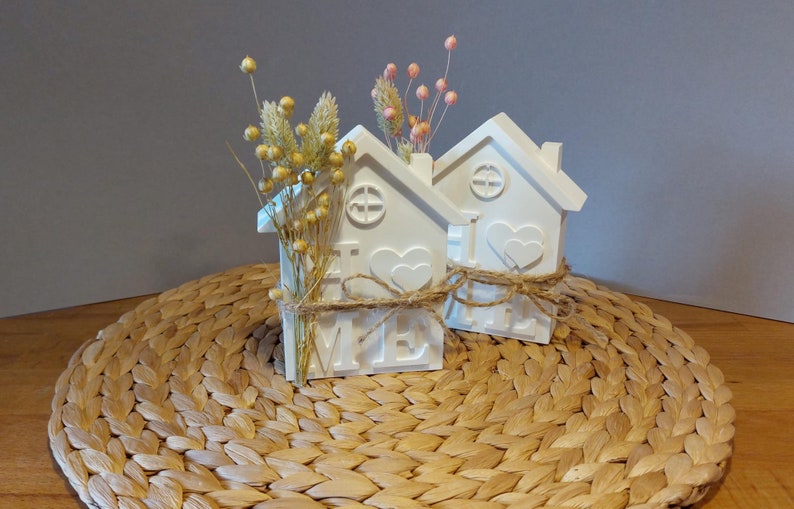 Könnte beinhalten: Zwei weiße, hausförmige Dekorationsvasen mit Trockenblumen. Jede Vase hat ein Herz und das Wort "HOME" auf der Vorderseite. Die Vasen sind mit Bindfaden gebunden und stehen auf einer gewebten Matte. Eine Vase hat gelbe, die andere rosa Blüten.