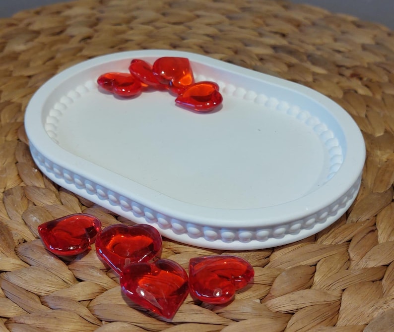 Könnte beinhalten: Eine weiße ovale Schale mit Perlenrand, die mehrere herzförmige Glasstücke in Rot enthält. Zusätzliche rote Herzen sind um die Schale herum verstreut. Die Schale steht auf einer gewebten, strukturierten Oberfläche.