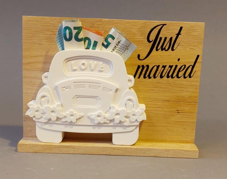 Könnte beinhalten: Ein weißer, autogeförmiger Hochzeitsgeschenkehalter mit dem Wort "LOVE" oben. Das Auto ist mit Blumen verziert und enthält gefaltete Banknoten. Der hölzerne Hintergrund trägt die Worte "Just married" in schwarzer Schrift.