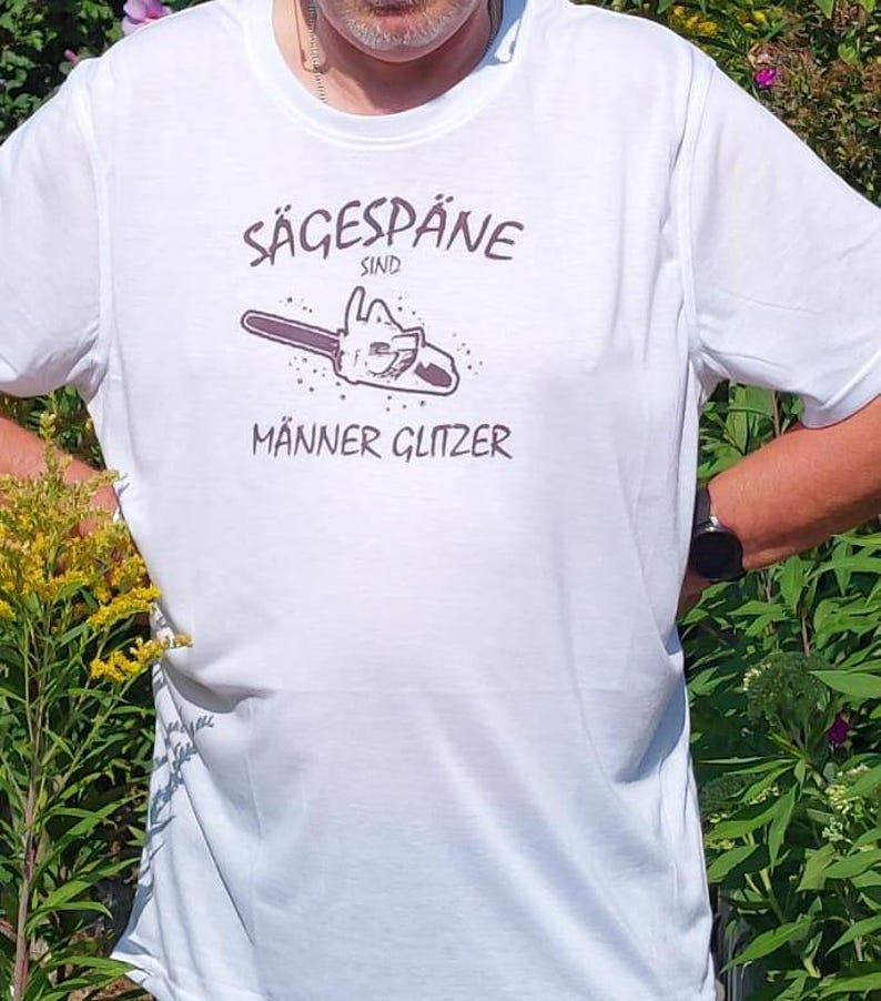 Könnte beinhalten: Weißes T-Shirt mit braunem Grafikdesign. Das Design zeigt eine Kettensägen-Illustration und den deutschen Text "Sägespäne sind Männer Glitzer". Das T-Shirt hat kurze Ärmel.