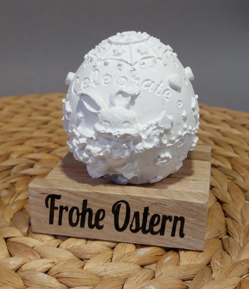 Könnte beinhalten: Weißes, eiförmiges Osterdekor mit dem Wort "Celebrate" und einem Hasen-Design. Die Dekoration steht auf einem Holzsockel mit dem Schriftzug "Frohe Ostern" in Schwarz.