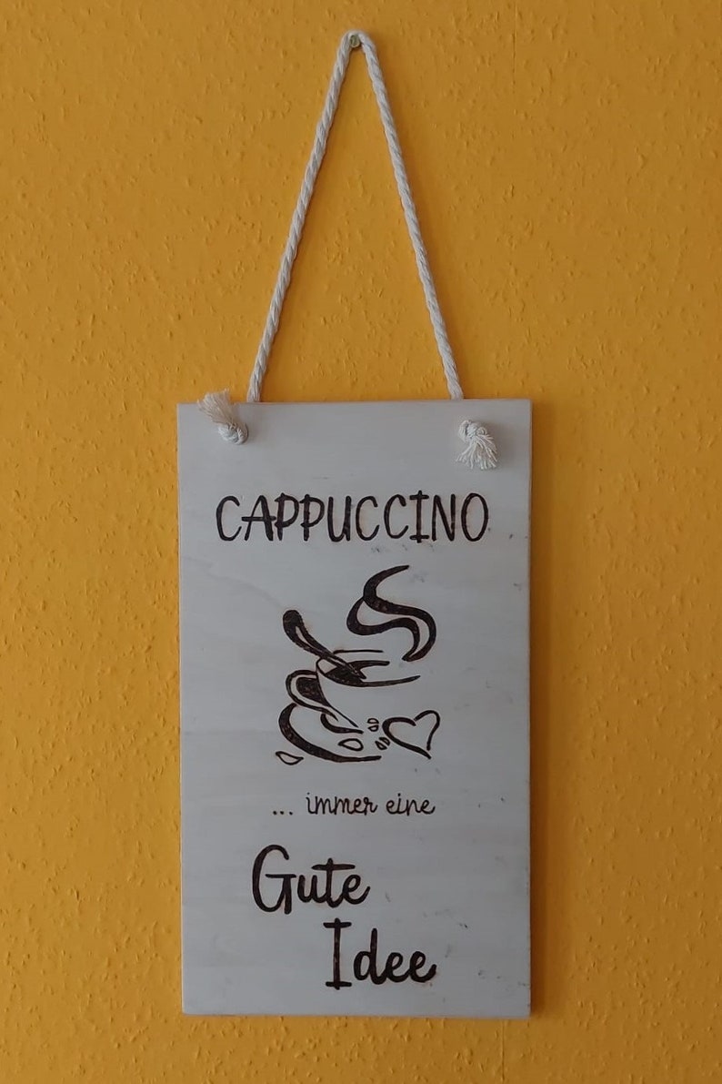 Könnte beinhalten: Ein weißes Holzschild mit einem Seilhalter. Das Schild hat eine schwarz-weiße Illustration einer Tasse Cappuccino mit aufsteigendem Dampf. Der Text auf dem Schild lautet "Cappuccino... immer eine gute Idee".