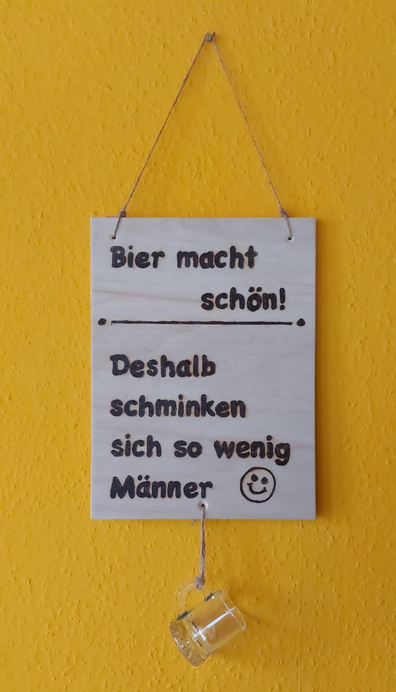Könnte beinhalten: Holzschild mit deutschem Text: "Bier macht schön! Deshalb schminken sich so wenig Männer." Darunter hängt ein kleiner Bierkrug aus Glas. Das Schild ist mit einer Schnur an einer gelben Wand befestigt.