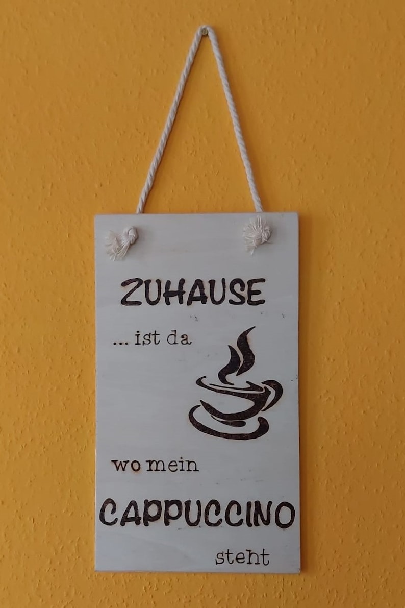 Könnte beinhalten: Ein weißes Holzschild mit dem Text "Zuhause... ist da wo mein Cappuccino steht" und einer schwarzen Silhouette einer Kaffeetasse mit aufsteigendem Dampf.