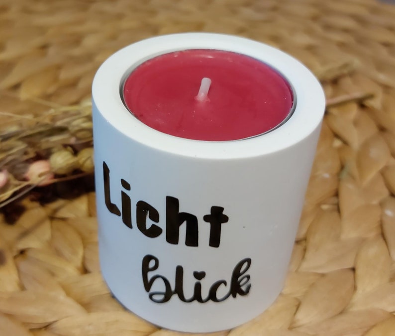 Könnte beinhalten: Weißer Kerzenhalter mit der Aufschrift "Licht Glück" in schwarzer Schrift. Der Halter hat eine rote Kerze drin.