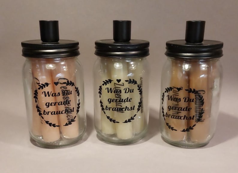 Könnte beinhalten: Drei Glasbehälter mit schwarzen Deckeln und Etiketten, auf denen "Was Du gerade brauchst" steht. Die Behälter sind mit kleinen, weißen und beigefarbenen Kerzen gefüllt.