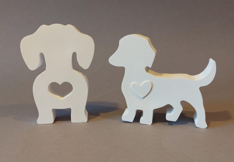 Könnte beinhalten: Zwei weiße Dackel-Figuren aus Holz. Ein Dackel steht mit eingerolltem Schwanz, der andere sitzt mit einem Herz-Ausschnitt im Hinterteil. Beide Figuren haben ein kleines Herz auf der Brust.
