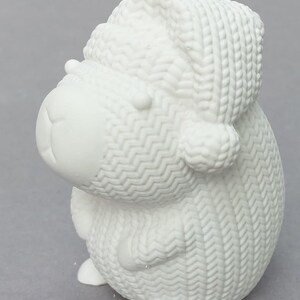 Könnte beinhalten: Eine weiße Keramik-Schaf-Figur mit gestrickter Mütze und Pullover. Das Schaf hat einen runden Körper, kleine Ohren und ein lächelndes Gesicht. Die Figur ist etwa 10 cm hoch.