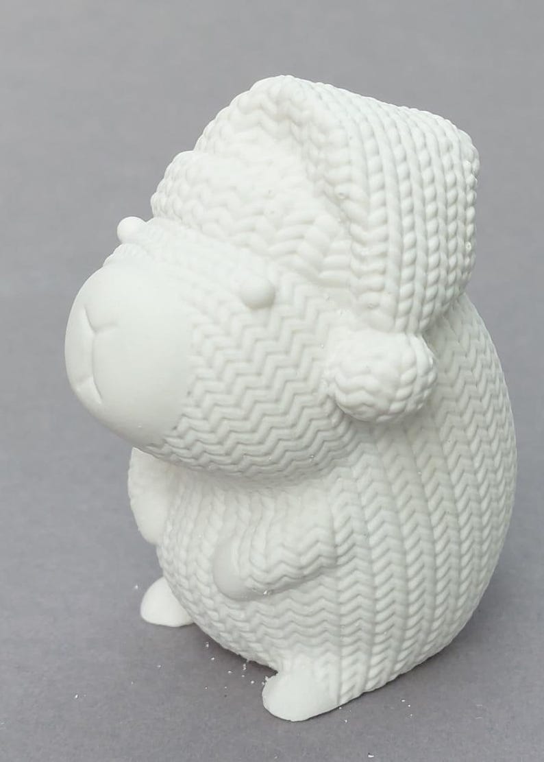 Könnte beinhalten: Eine weiße Keramik-Schaf-Figur mit gestrickter Mütze und Pullover. Das Schaf hat einen runden Körper, kleine Ohren und ein lächelndes Gesicht. Die Figur ist etwa 10 cm hoch.