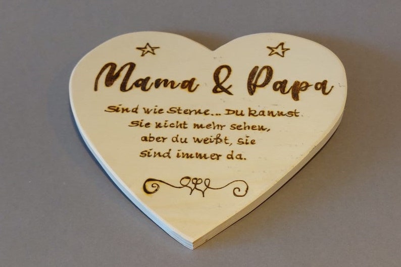 Könnte beinhalten: Ein weißes herzförmiges Holzschild mit dem Text "Mama & Papa" und einer Botschaft über Sterne. Der Text lautet: "Sind wie Sterne... Du kannst. Sie nicht mehr sehen, aber du weißt, sie sind immer da."