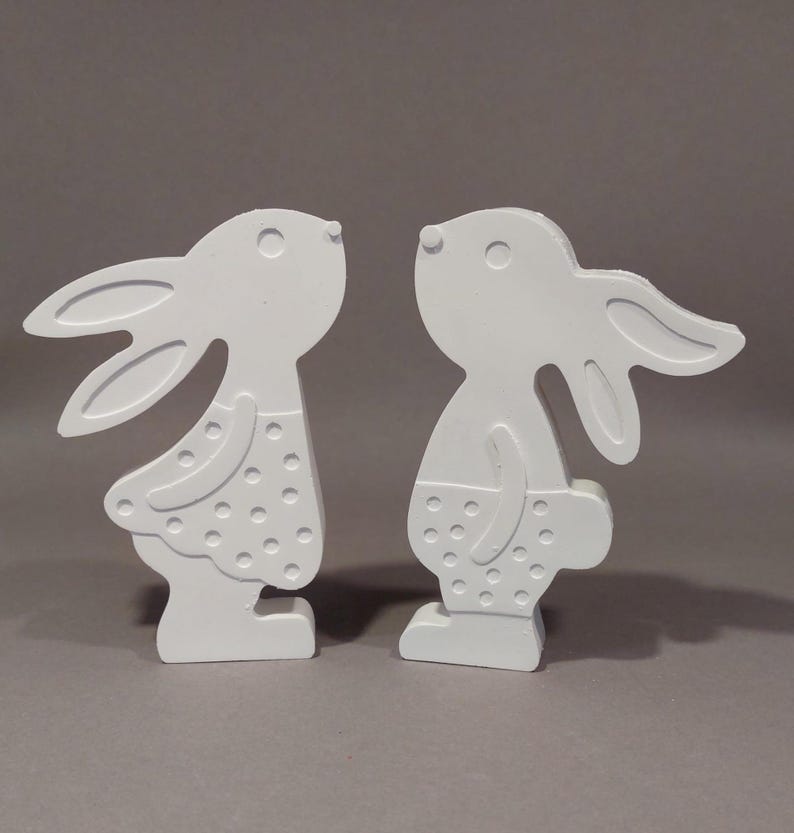 Könnte beinhalten: Zwei weiße Holzhasenfiguren. Jeder Hase hat große Ohren, einen runden Körper und trägt ein gepunktetes Outfit. Die Hasen stehen sich gegenüber und schaffen eine charmante und skurrile Darstellung.