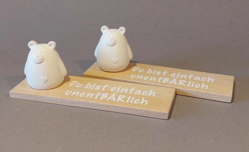 Könnte beinhalten: Zwei weiße Keramik-Bärenfiguren mit Herzen auf der Brust, die jeweils auf einem Holzsockel mit dem Text "Du bist einfach unentbeärlich" (Du bist einfach unverzichtbar) in weißer Schrift stehen.