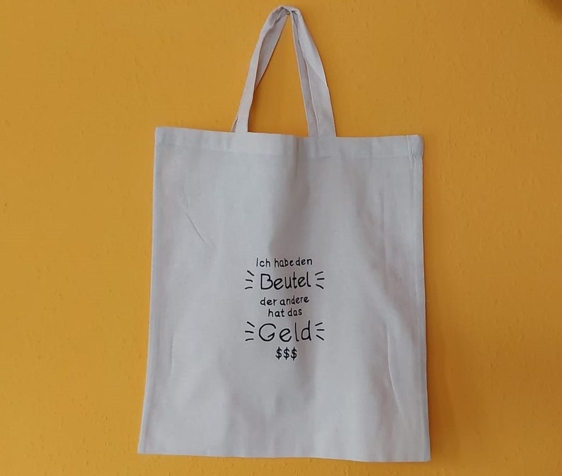 Könnte beinhalten: Weiße Stofftasche mit schwarzem Text, der "Ich habe den Beutel der andere hat das Geld $$$" lautet.