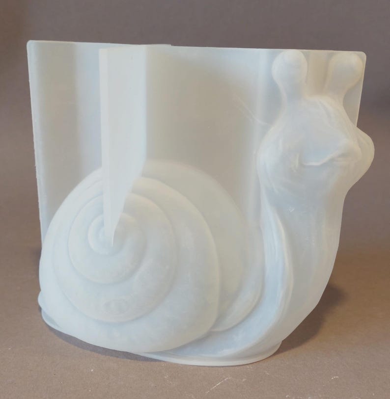 Könnte beinhalten: Ein weißer, transluzenter Keramik-Pflanztopf mit Schnecken-Design. Das Schneckenhaus ist spiralförmig, und Körper und Kopf sind detailliert. Der Pflanztopf hat eine rechteckige Form mit einer geschwungenen Vorderseite.