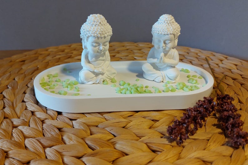 Könnte beinhalten: Eine weiße, ovale Dekoschale mit zwei kleinen weißen Buddha-Statuen. Die Statuen sitzen in meditativer Pose. Die Schale ist mit kleinen grünen Steinen verziert. Der Hintergrund ist eine gewebte Matte.