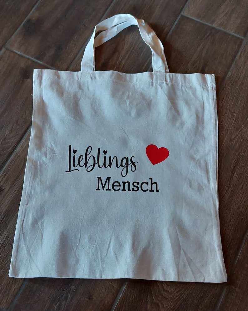 Könnte beinhalten: Weiße Stofftasche mit schwarzem Text "Lieblings Mensch" und einem roten Herz.
