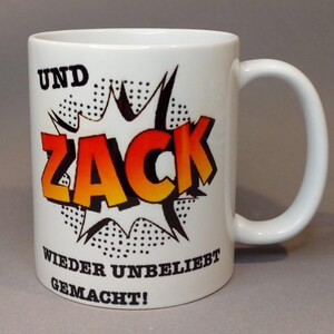 Könnte beinhalten: Weiße Keramiktasse mit einer schwarz-weißen Comic-Grafik mit dem Wort "ZACK" in Rot und Gelb. Der Text "UND ZACK WIEDER UNBELIEBT GEMACHT!" ist auf die Tasse gedruckt.