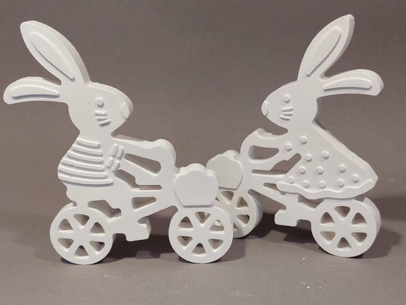 Könnte beinhalten: Zwei weiße Holzhasen-Figuren auf Fahrrädern. Die Hasen haben detaillierte Merkmale, einschließlich langer Ohren und Gesichtsausdrücke. Einer trägt ein gestreiftes Hemd, der andere ein gepunktetes Kleid. Die Fahrräder haben detaillierte Räder.