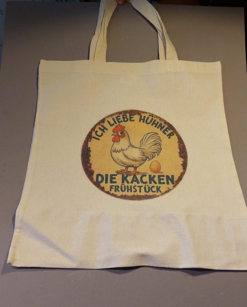 Könnte beinhalten: Cremefarbene Tragetasche mit einem kreisförmigen Aufdruck, der ein Cartoon-Huhn und den deutschen Text "Ich Liebe Hühner Die Kacken Frühstück" zeigt. Das Huhn ist braun, rot und weiß, mit einem gelben Ei. Die Tasche hat zwei Griffe.