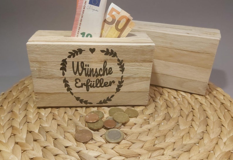 Könnte beinhalten: Holzkiste mit einem Schlitz für Geld, gefüllt mit Euro-Banknoten. Die Kiste ist mit den Worten "Wünsche Erfüller" in einem dekorativen Kranz graviert. Mehrere Münzen liegen vor der Kiste. Eine zweite, ähnliche Kiste ist sichtbar.