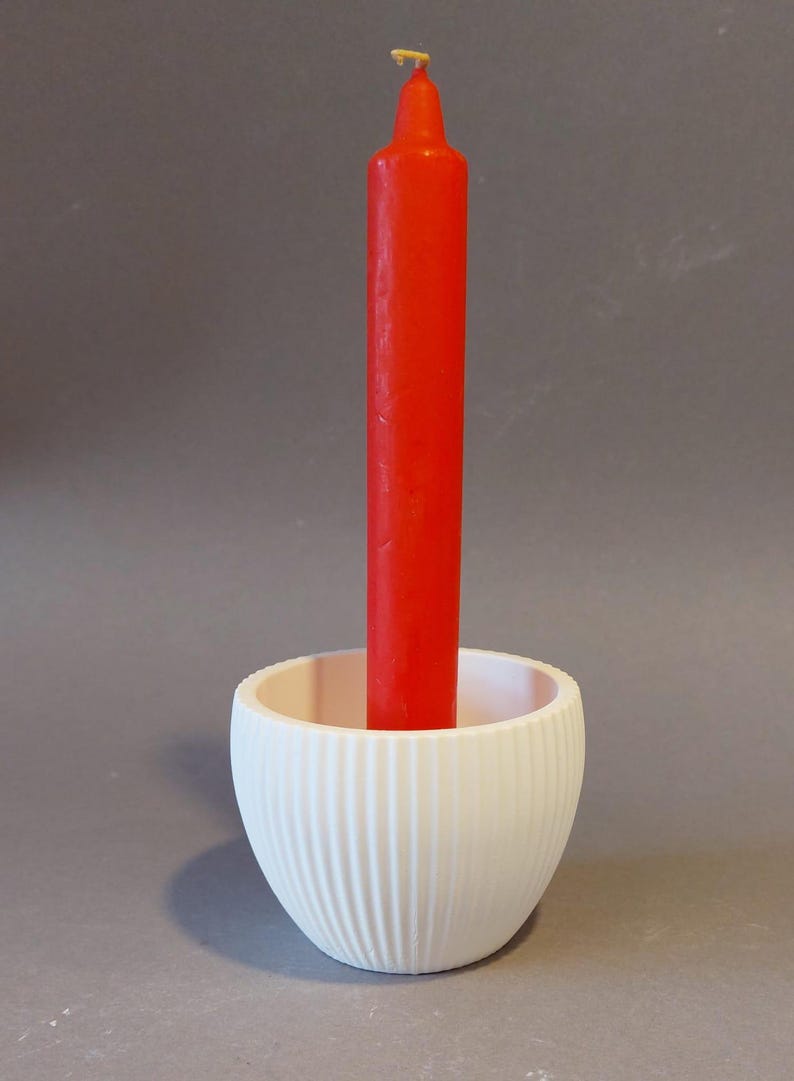 Könnte beinhalten: Eine rote Kerze in einem weißen, geriffelten Kerzenhalter. Die Kerze ist mittig in dem Halter platziert, der eine abgerundete Form hat. Der Kerzenhalter besteht aus einem glatten, matten Material.