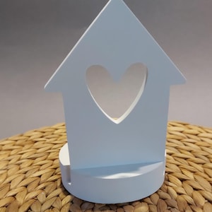 Könnte beinhalten: Weißer, hölzerner Kerzenhalter in Form eines Hauses mit einem ausgeschnittenen Herzen. Der Halter steht auf einer geflochtenen Weidenoberfläche.