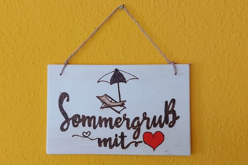 Könnte beinhalten: Ein weißes Holzschild mit einer schwarz-weißen Zeichnung eines Sonnenschirms und eines Liegestuhls. Das Schild sagt "Sommergrus mit" mit einem roten Herzen.