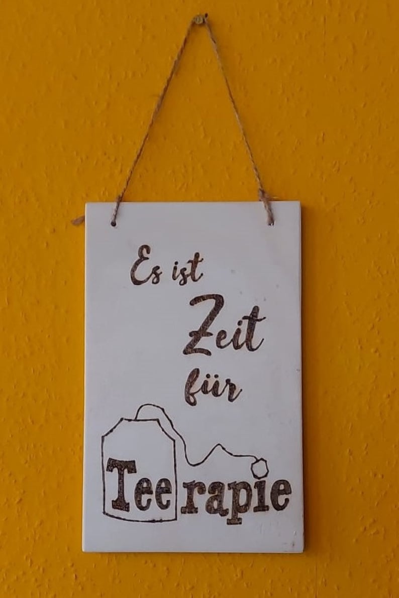 Könnte beinhalten: Ein weißes Holzschild mit einer braunen Teebetrauchillustration und dem Text "Es ist Zeit für Teerapie". Das Schild hängt an einer Schnur.