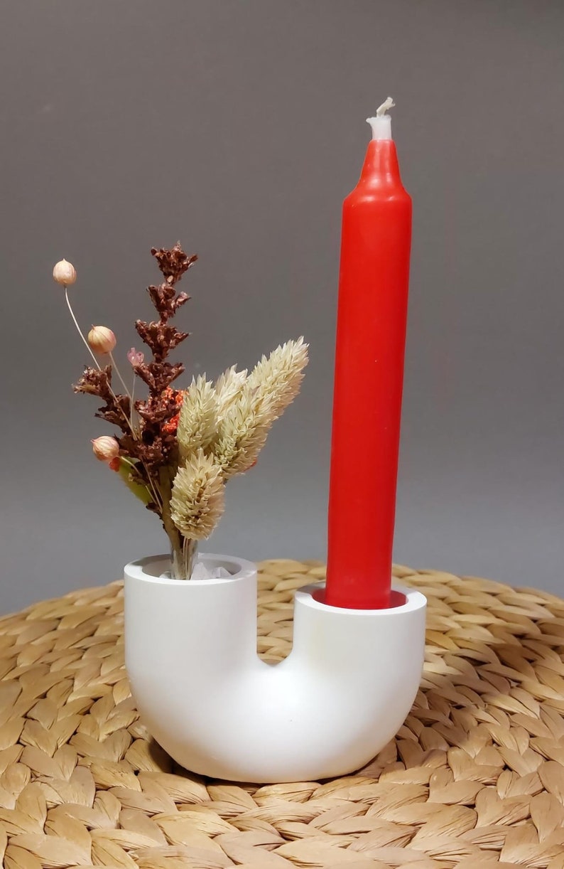 Könnte beinhalten: Ein weißer, U-förmiger Kerzenhalter mit einer roten Kerze und einem Trockenblumenarrangement. Die Kerze ist leuchtend rot, und die Blumen sind eine Mischung aus Beige, Braun und Rosa. Der Halter steht auf einer gewebten Matte.