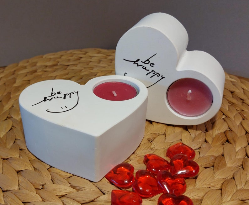 Könnte beinhalten: Zwei weiße, herzförmige Kerzenhalter aus Keramik mit der Aufschrift "be happy". Jeder Halter hat eine kleine, runde, rote Kerze darin. Die Halter stehen auf einer geflochtenen Weidenoberfläche mit roten herzförmigen Dekorationen.