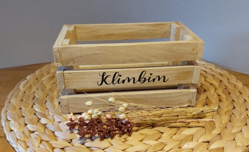 Könnte beinhalten: Eine kleine Holzkiste mit dem Wort "Klimbim" auf der Vorderseite. Die Kiste steht auf einer geflochtenen Strohmatte.