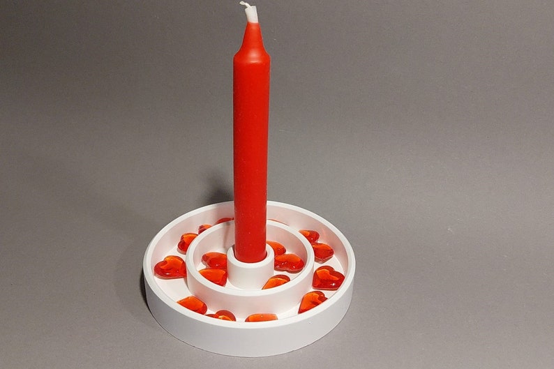 Könnte beinhalten: Ein weißer Kerzenhalter mit zwei Ebenen, verziert mit roten herzförmigen Akzenten. Eine einzelne rote Kerze sitzt in der Mitte.