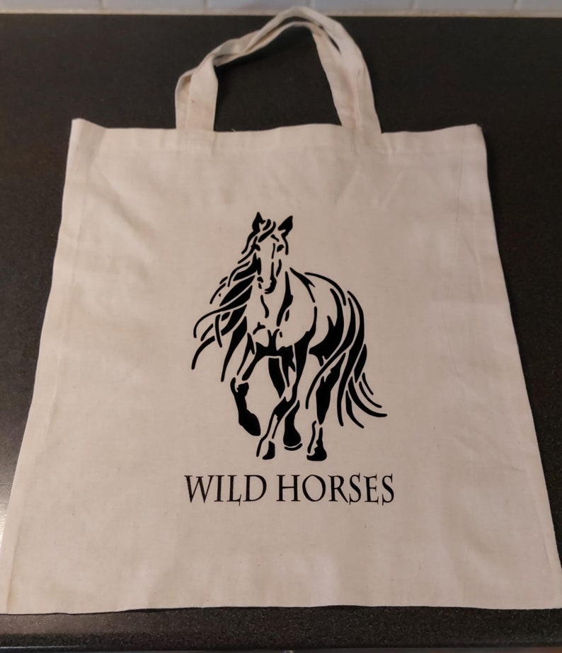 Könnte beinhalten: Eine weiße Stofftasche mit schwarzen Henkeln. Die Tasche zeigt eine schwarze Silhouette eines Pferdes mit einer fließenden Mähne und einem Schweif. Der Text "WILD HORSES" ist in Schwarz unter dem Pferd aufgedruckt.