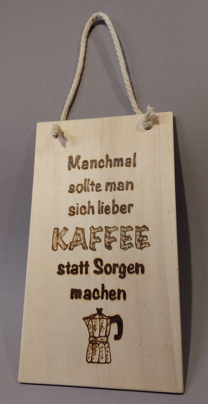 Könnte beinhalten: Ein Holzschild mit einem Seil zum Aufhängen, mit dem Text "Manchmal sollte man sich lieber Kaffee statt Sorgen machen" und einer Zeichnung einer Kaffeekanne.