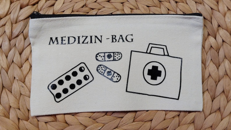 Könnte beinhalten: Eine weiße Stofftasche mit schwarzem Reißverschluss und dem aufgedruckten Text "MEDIZIN-BAG" auf der Vorderseite. Die Tasche zeigt schwarze Illustrationen einer Pillendose, zweier Bandagen und einer Erste-Hilfe-Box.