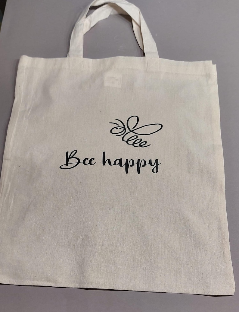 Könnte beinhalten: Eine weiße Stofftasche mit schwarzem Text, der "Bee happy" lautet, und einer schwarzen Bienenillustration.