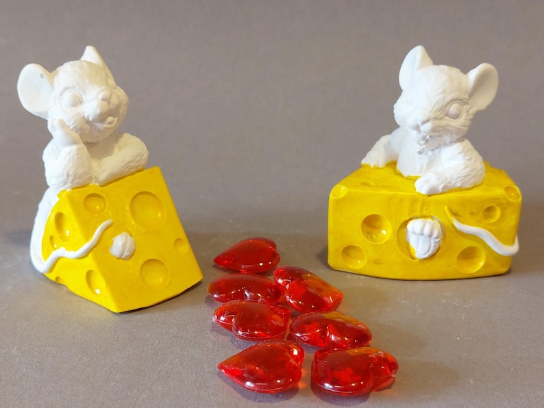 Könnte beinhalten: Zwei weiße Mäusefiguren sitzen auf gelben Käseblöcken mit Löchern. Rote herzförmige Glasperlen sind vor dem Käse und den Mäusen verstreut. Die Käseblöcke haben weiße Akzente.
