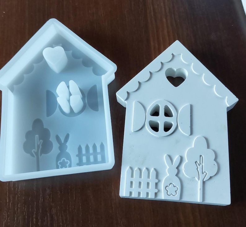 Könnte beinhalten: Zwei weiße Silikonformen in Form von Häusern. Eine Form hat ein Herz, einen Schmetterling, einen Baum, ein Kaninchen und ein Zaun-Design. Die andere Form hat ein herzförmiges Fenster, ein rundes Fenster, ein Kaninchen, einen Baum und ein Zaun-Design.