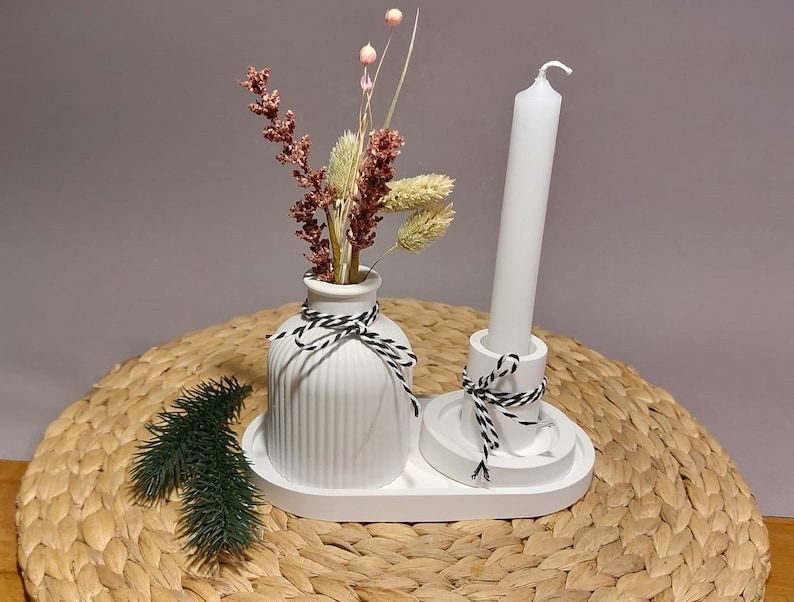 Könnte beinhalten: Weiße Keramikvase mit einem schwarz-weißen Band, ein weißer Kerzenhalter mit einem schwarz-weißen Band und eine weiße Kerze auf einer geflochtenen Weidenoberfläche.