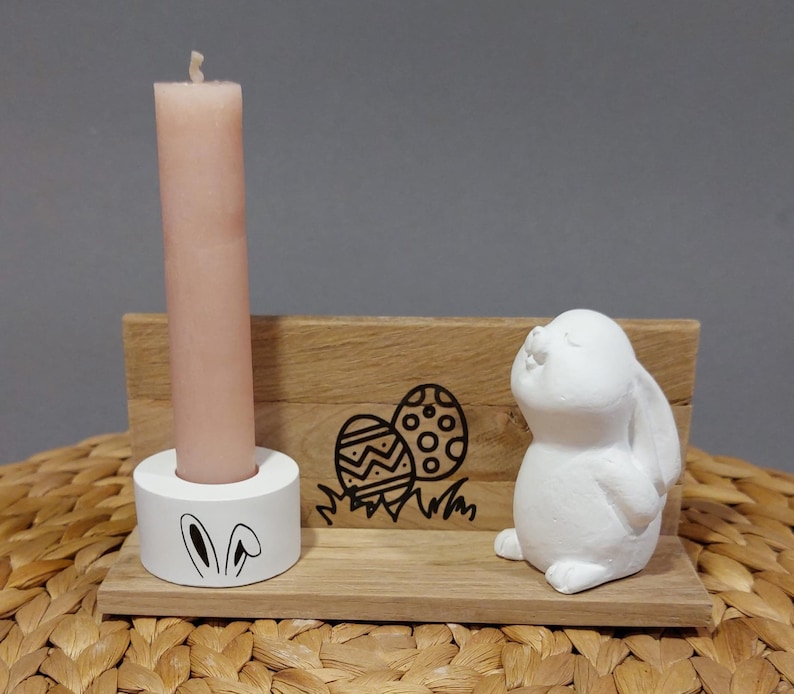 Könnte beinhalten: Eine Osterdekoration aus Holz mit einer rosa Kerze in einem weißen Halter mit Hasenohren, einer weißen Hasenfigur und einem Holz-Hintergrund mit Osterei-Grafiken. Die Dekoration steht auf einer gewebten Oberfläche.