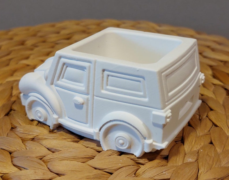 Könnte beinhalten: Weißer Keramik-Pflanztopf in Form eines Oldtimers. Der Topf hat eine quadratische Öffnung oben zum Pflanzen.