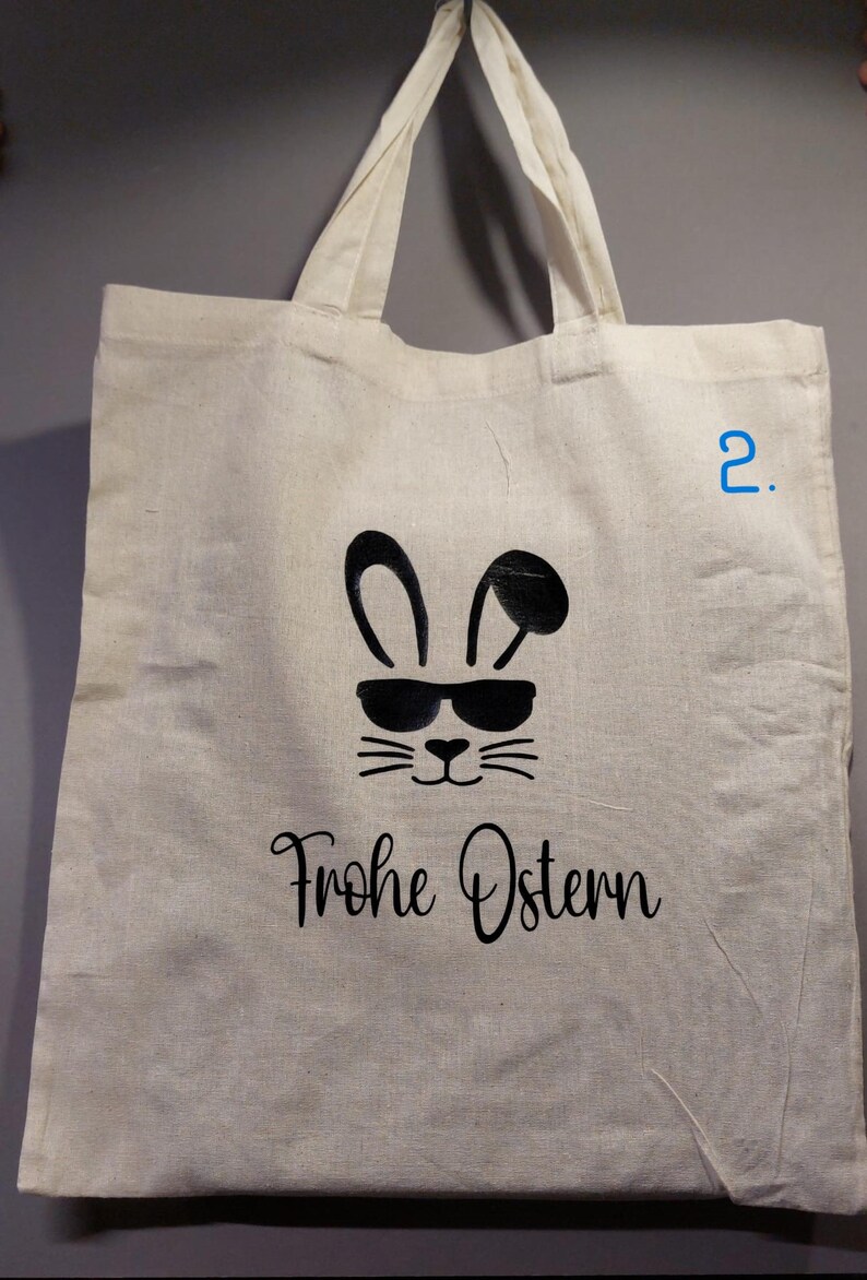 Könnte beinhalten: Eine cremefarbene Tragetasche mit schwarzen Grafiken. Das Design zeigt ein Hasengesicht mit Sonnenbrille und die Worte "Frohe Ostern" in einer Schriftart. Die Tasche hat zwei Griffe.